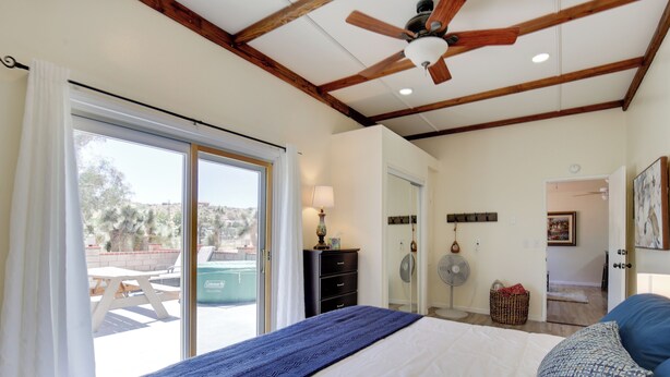 Une chambre à coucher avec un lit, un ventilateur de plafond, une fenêtre avec des rideaux, une vue sur l’extérieur et une porte donnant sur une autre pièce.