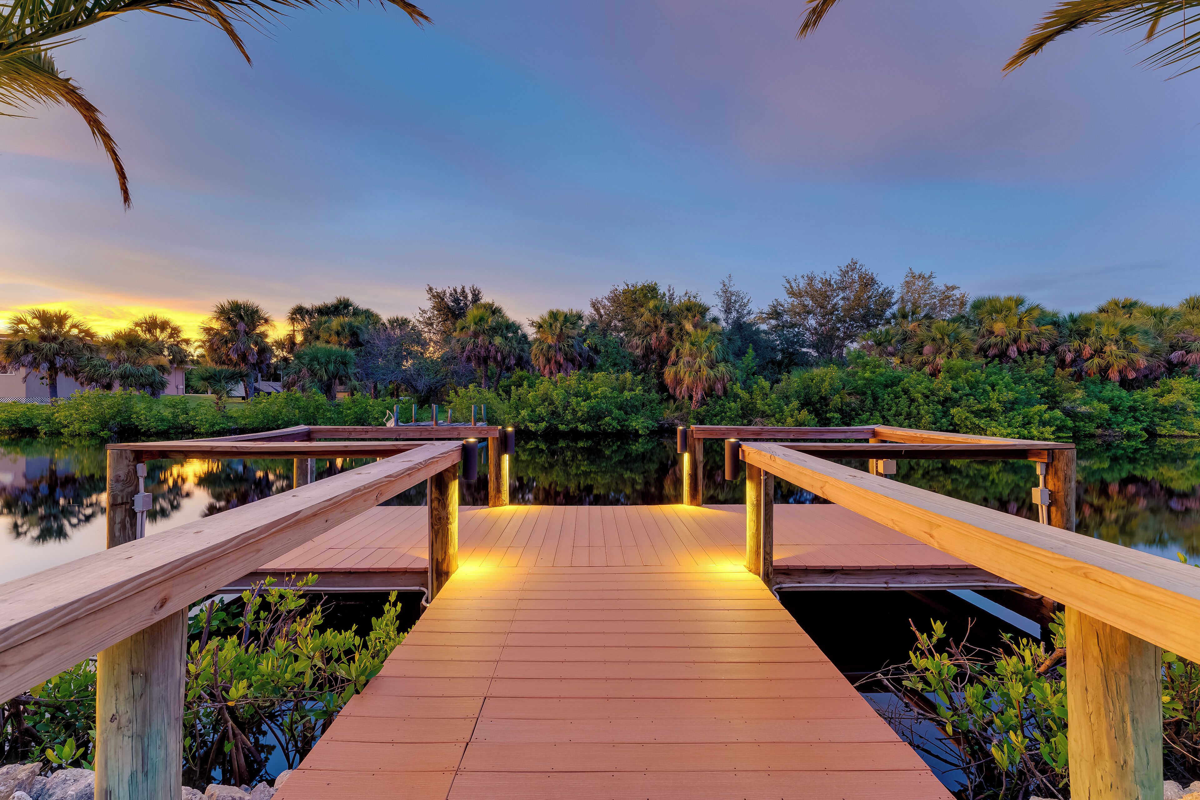 A wooden dock leading to a calm body of water, surrounded by lush greenery and palm trees.