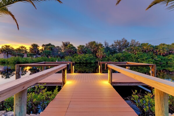 A wooden dock leading to a calm body of water, surrounded by lush greenery and palm trees.