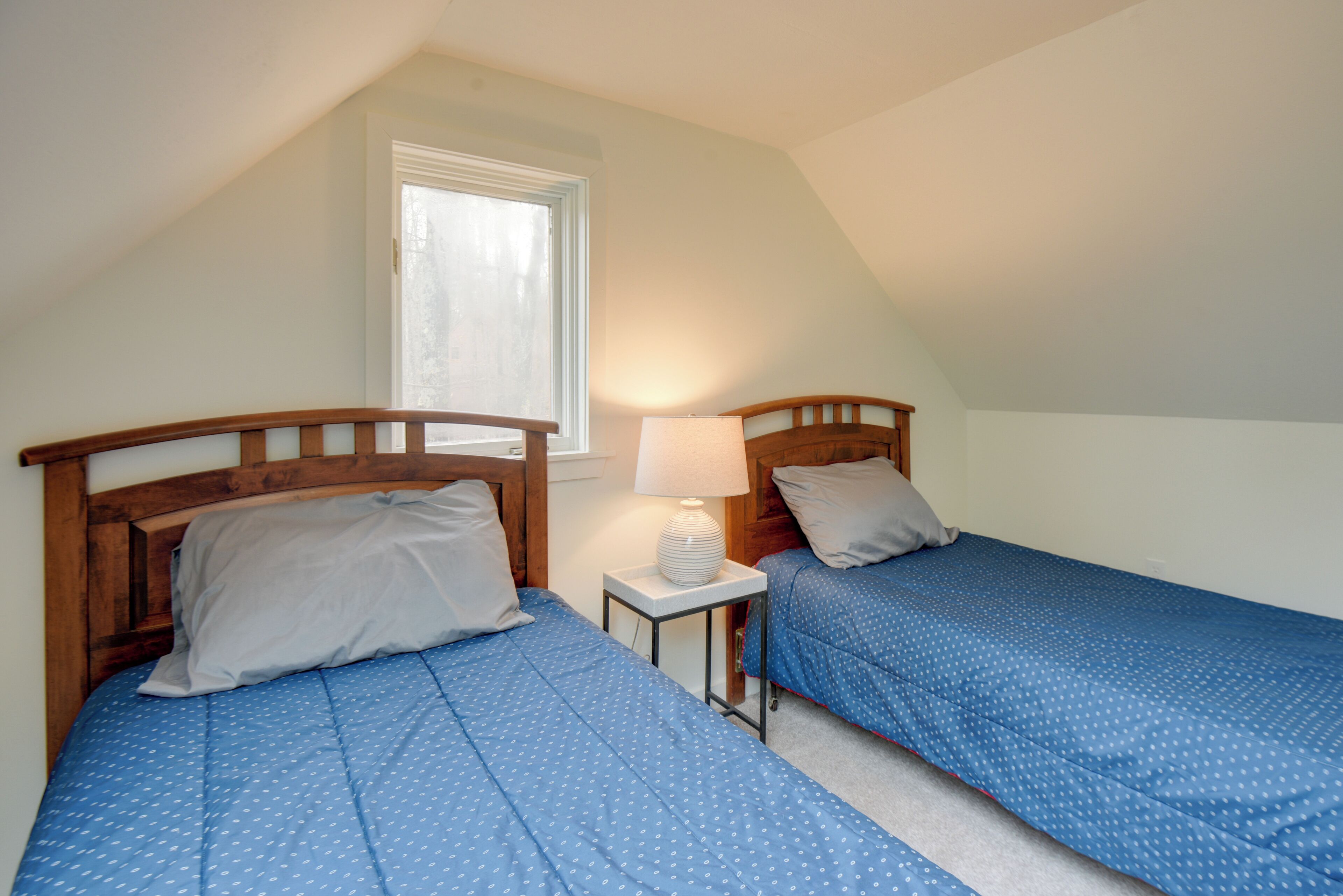 Quarto com duas camas de solteiro, mesa de cabeceira com luminária e janela.