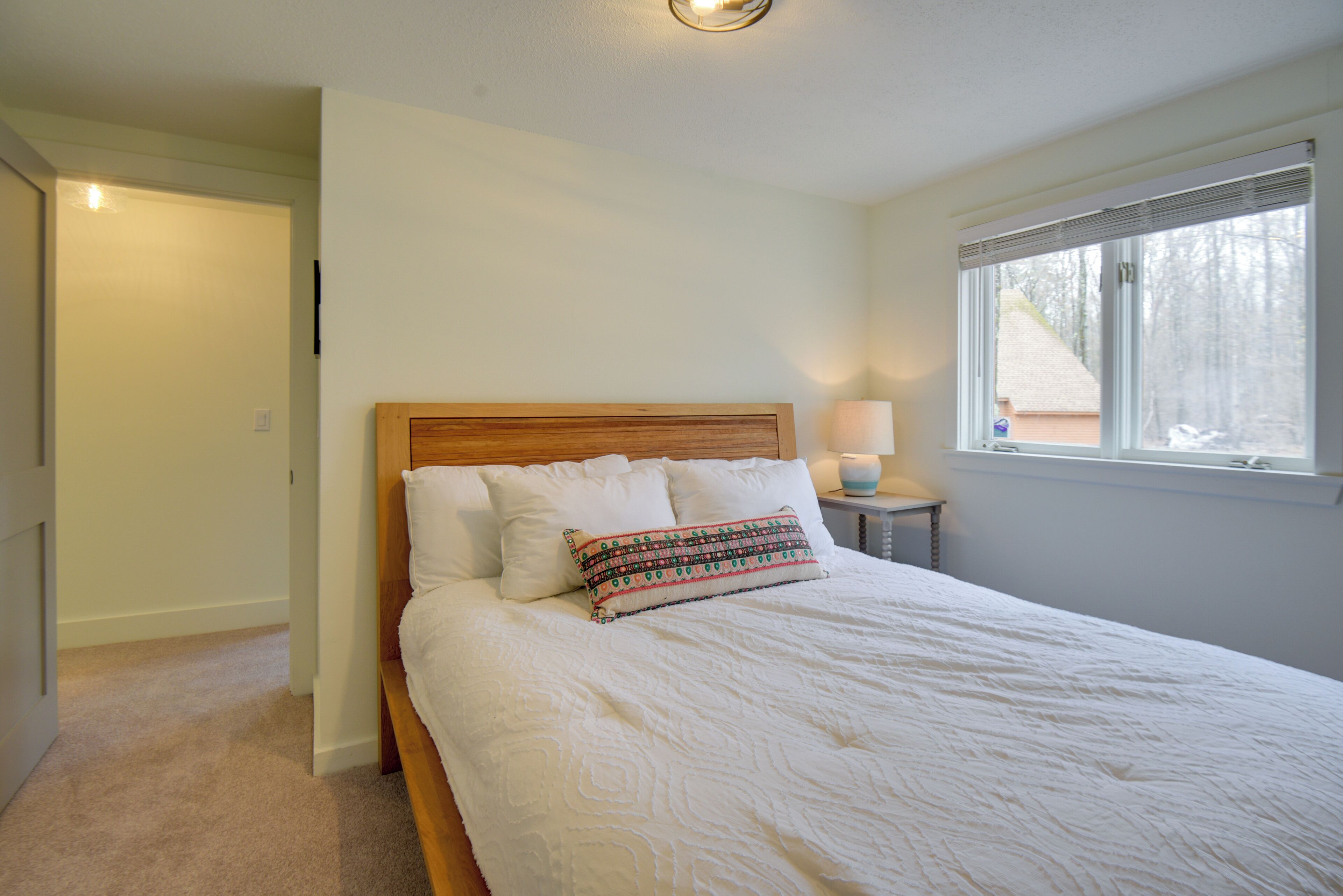 A neatly made bed with white bedding and a patterned pillow, a bedside lamp, and a window with blinds.