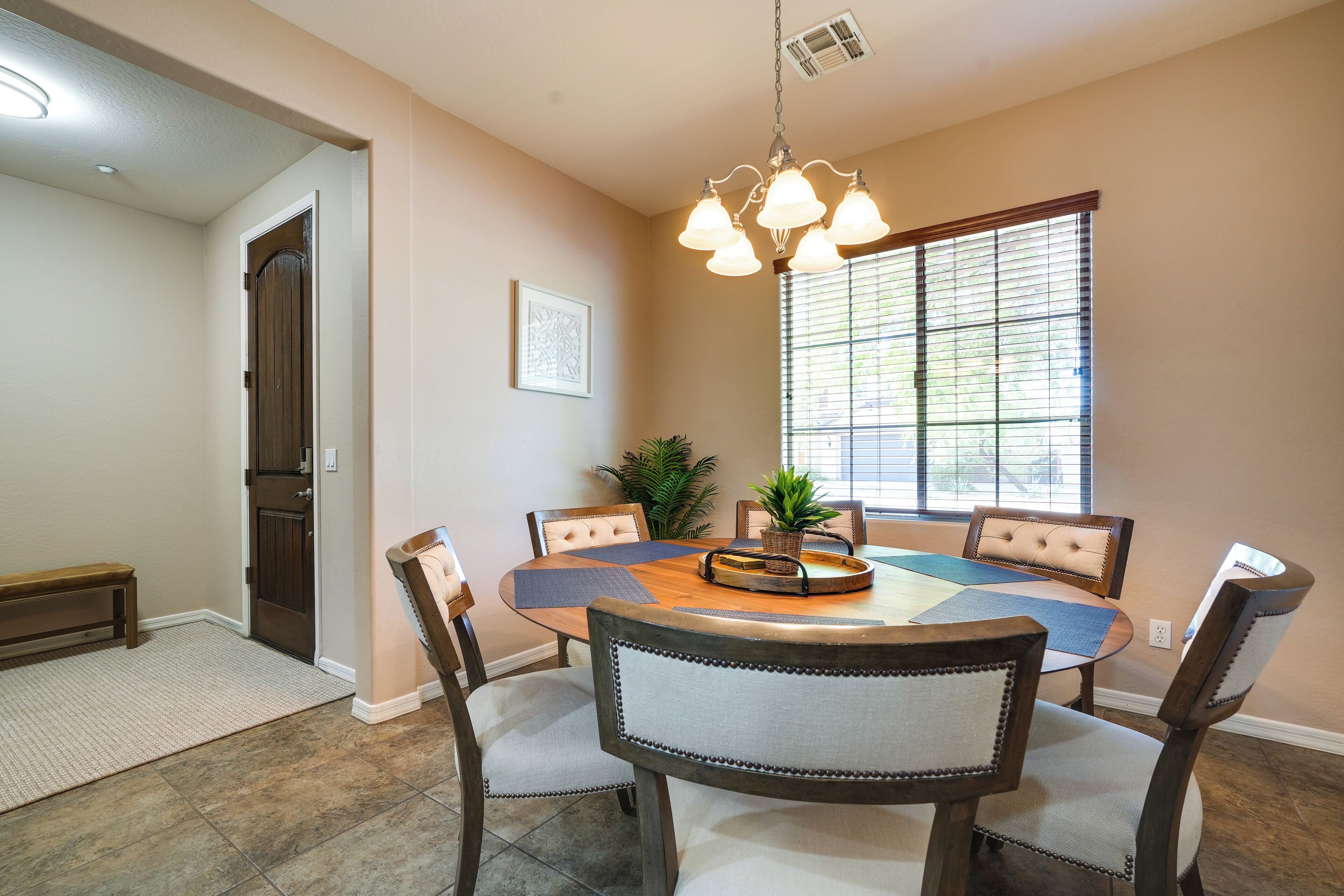 A dining area with a round table, six chairs, a chandelier, a window with blinds, and a door.