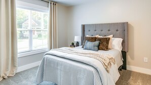 Une chambre avec un grand lit, une fenêtre avec des rideaux, une table de chevet avec une lampe, et une vue sur une maison à l’extérieur.