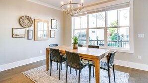 Une salle à manger avec une table en bois, six chaises, un lustre et de grandes fenêtres.