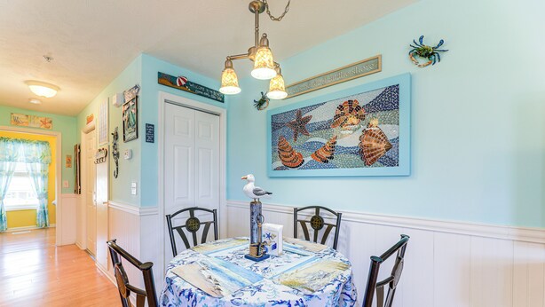 Un área de comedor con una mesa preparada para cuatro personas, rodeada de sillas, un centro de mesa decorativo y una pared con un cuadro de temática náutica.