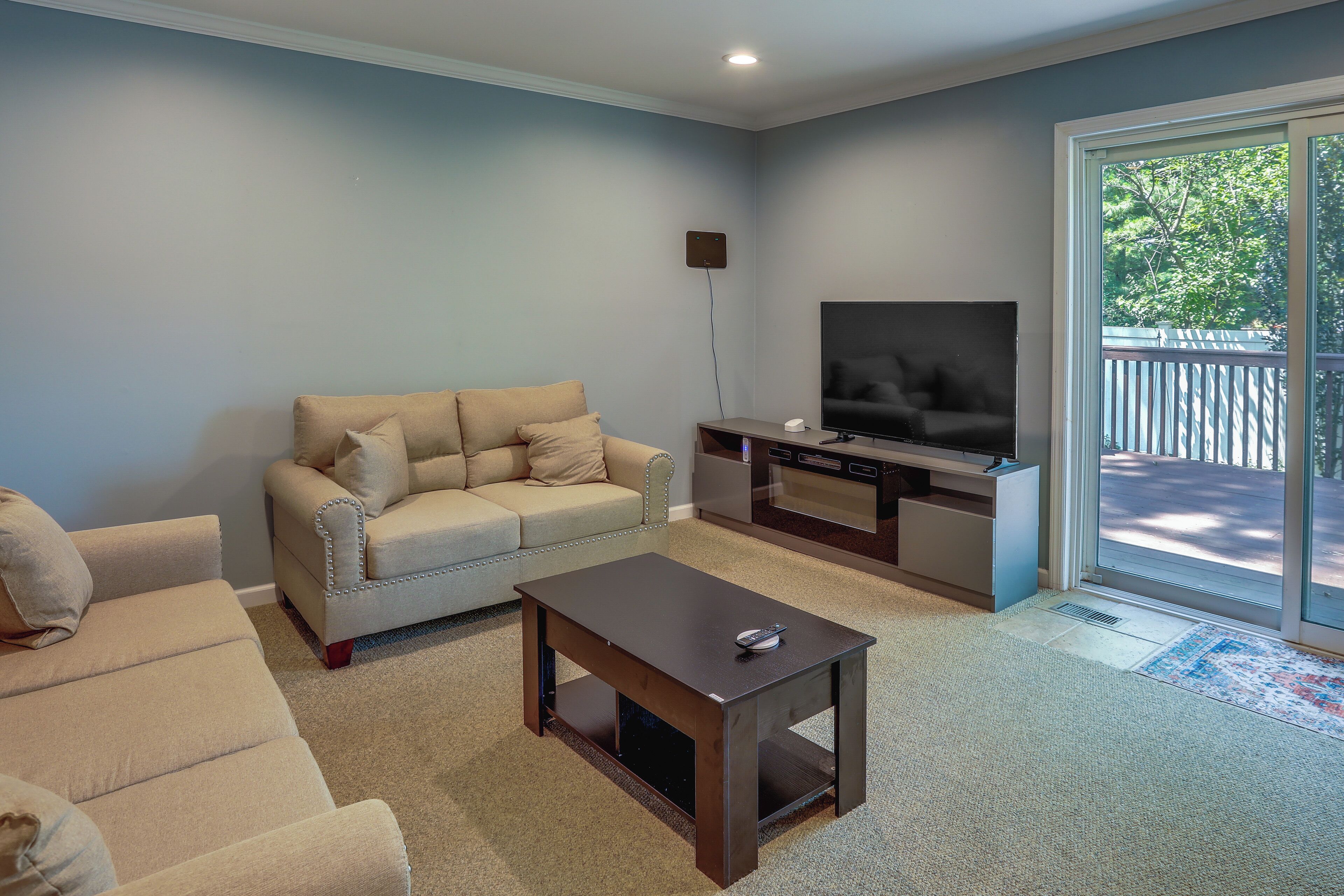 A living room with a beige sofa, a coffee table, a flat-screen TV, and a sliding glass door leading to a balcony.