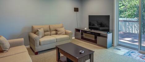 A living room with a beige sofa, a coffee table, a flat-screen TV, and a sliding glass door leading to a balcony.