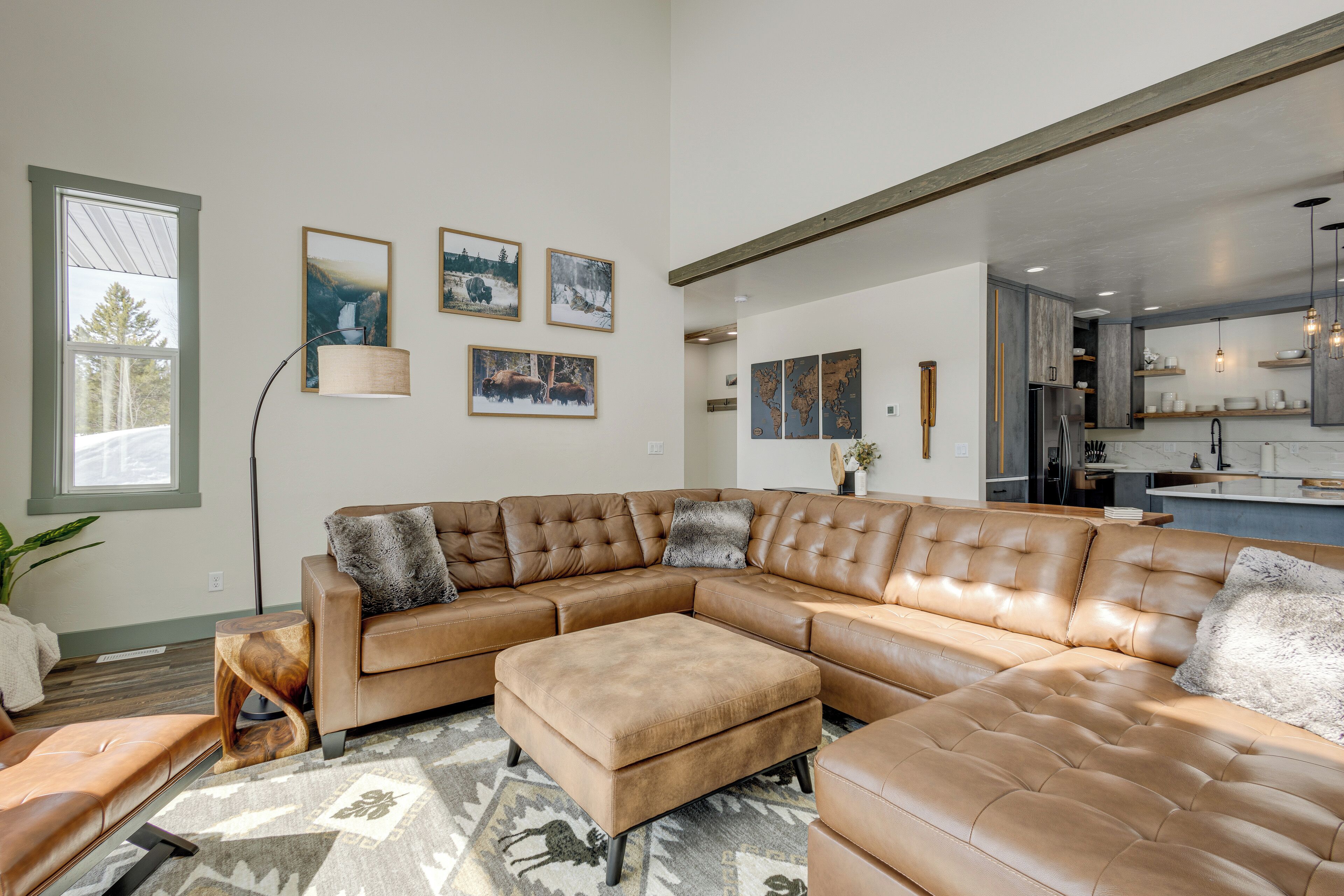 Un salon moderne avec un grand canapé d’angle, une table basse et un lampadaire. Des tableaux encadrés ornent le mur, et on aperçoit l’espace cuisine.