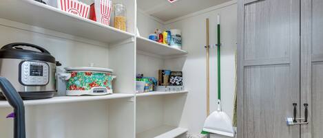 A kitchen with a microwave, a slow cooker, and a coffee maker on the counter. There are shelves above holding various items like cereal boxes and a mop.