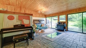 A wooden interior with a piano, a sofa, and a large window offering a view of greenery. - Catskills Mountain Cabin in Bloomville! (Bloomville)