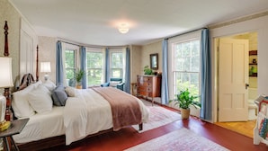 Een ruime slaapkamer met een groot bed, meerdere ramen, een dressoir en een deur naar een andere kamer.