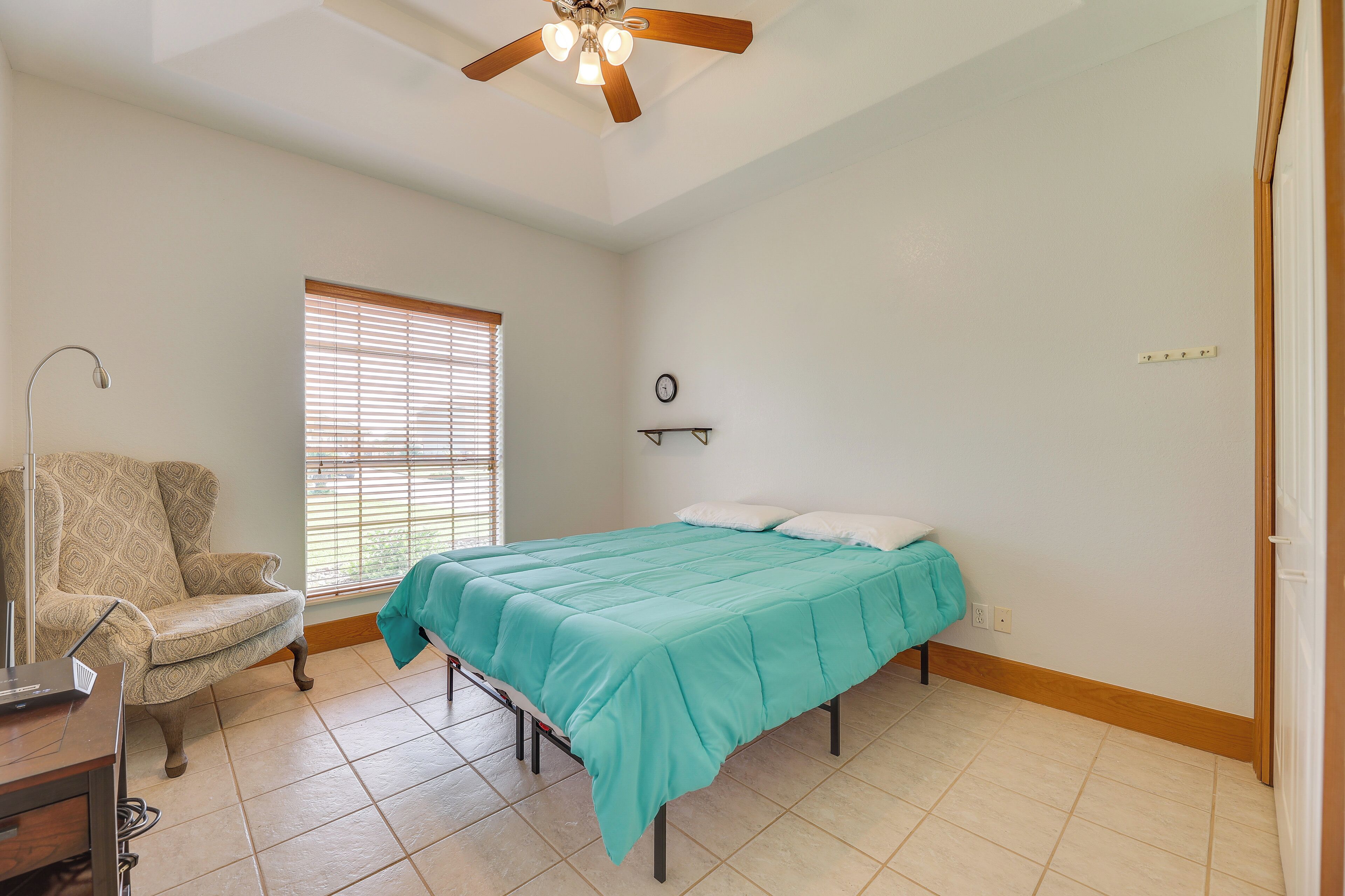 A bedroom with a bed, a chair, a ceiling fan, a window with blinds, and a wall clock.