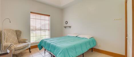 A bedroom with a bed, a chair, a ceiling fan, a window with blinds, and a wall clock.
