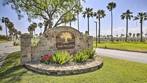 A stone sign with a tree and palm trees in the background, surrounded by grass and red flowers. - Swim, Golf & Unwind: Laguna Vista Vacation Home! (Port Isabel)