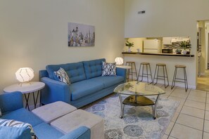 Un salon avec un canapé bleu, une table basse en verre et un coin cuisine comprenant un bar et des tabourets.