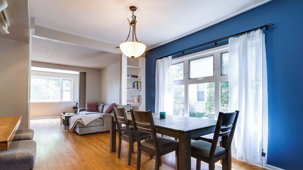 A dining area with a wooden table and chairs, a bookshelf, and a window with curtains.