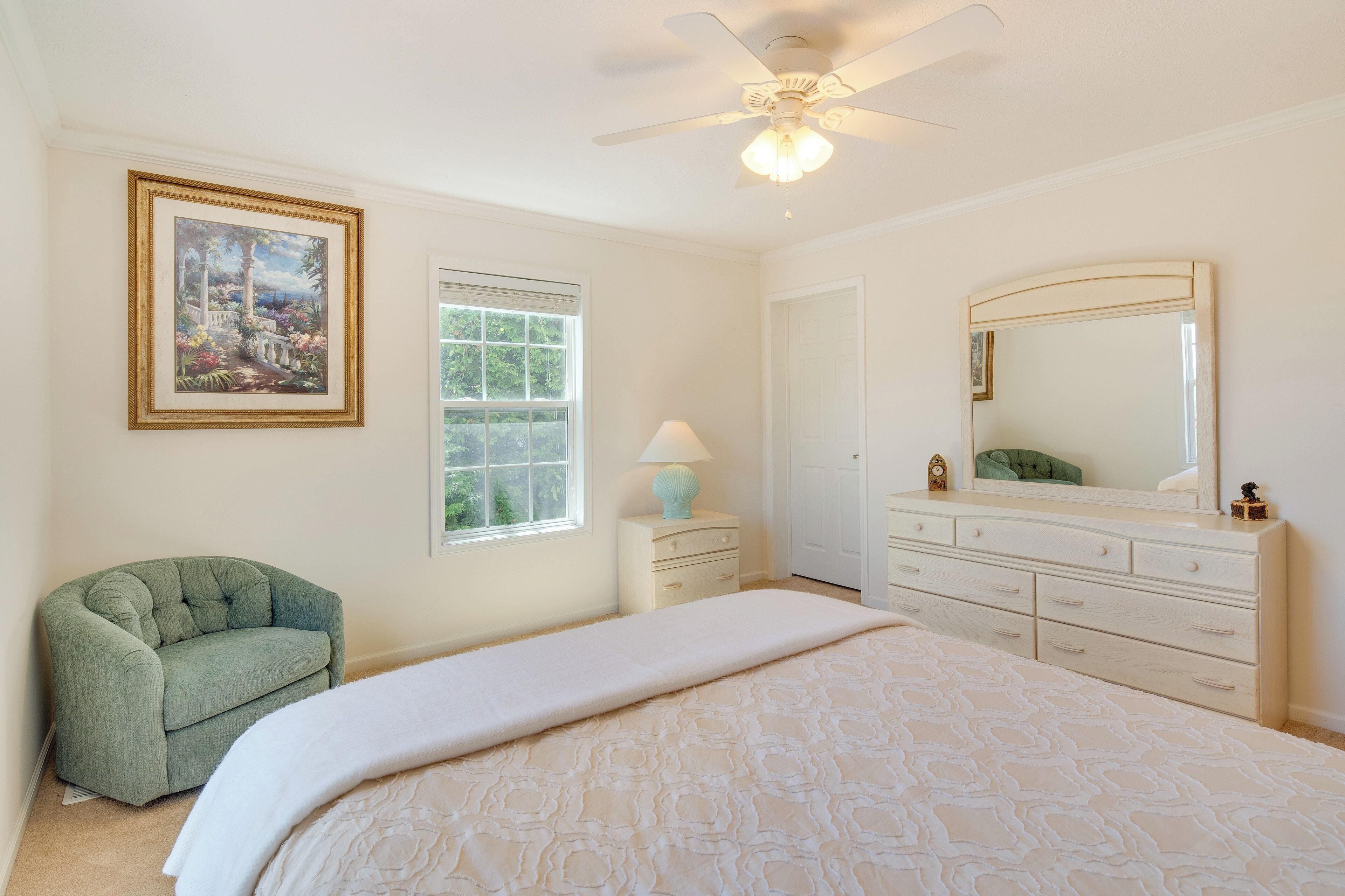 A bedroom with a bed, a ceiling fan, a dresser, a chair, a window, and a framed picture on the wall.