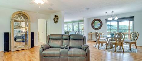 A spacious living room with a leather sofa, wooden dining table, and chairs, a ceiling fan, and large windows.