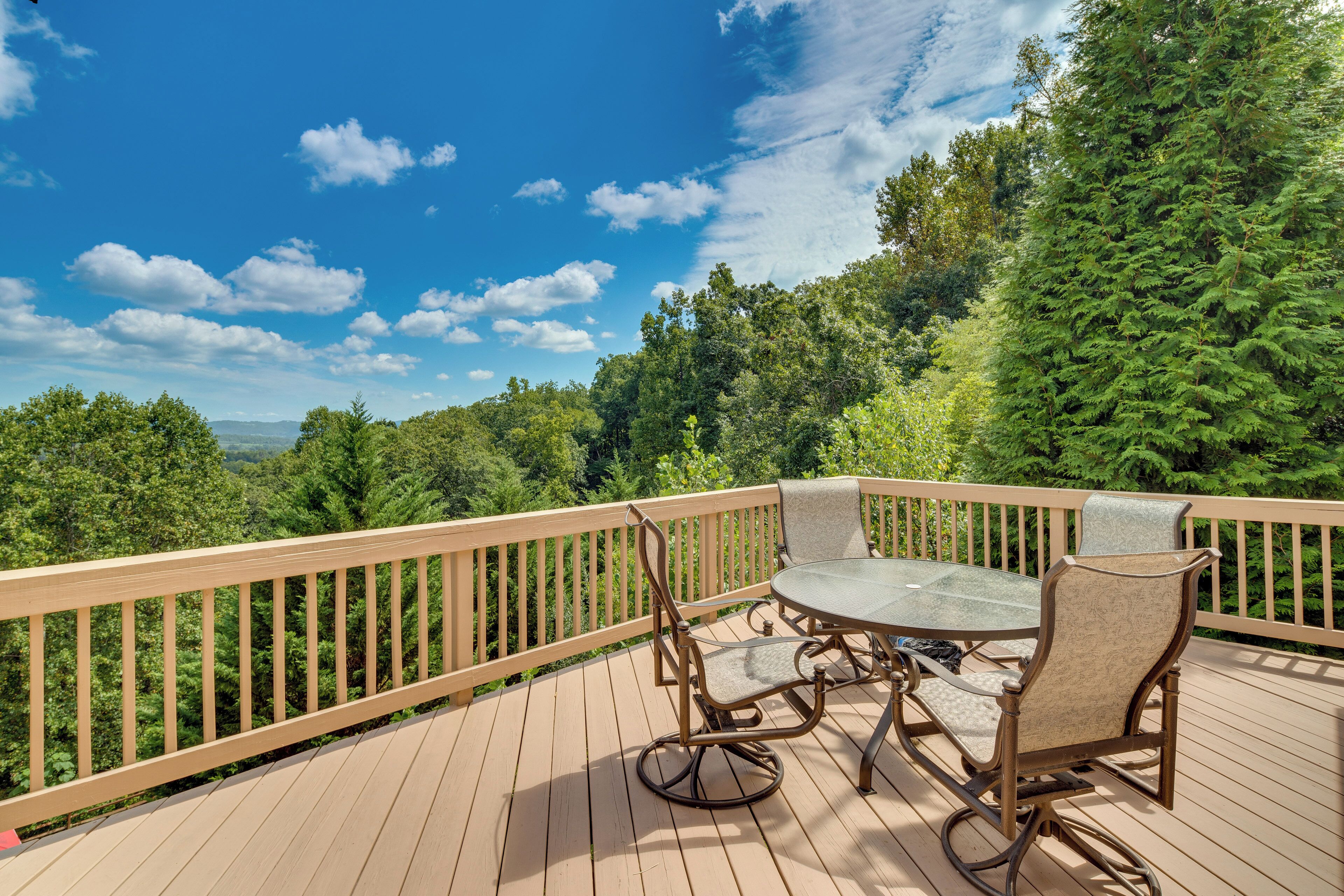 A wooden deck with a glass table and chairs, surrounded by a wooden railing, overlooking a forested area.