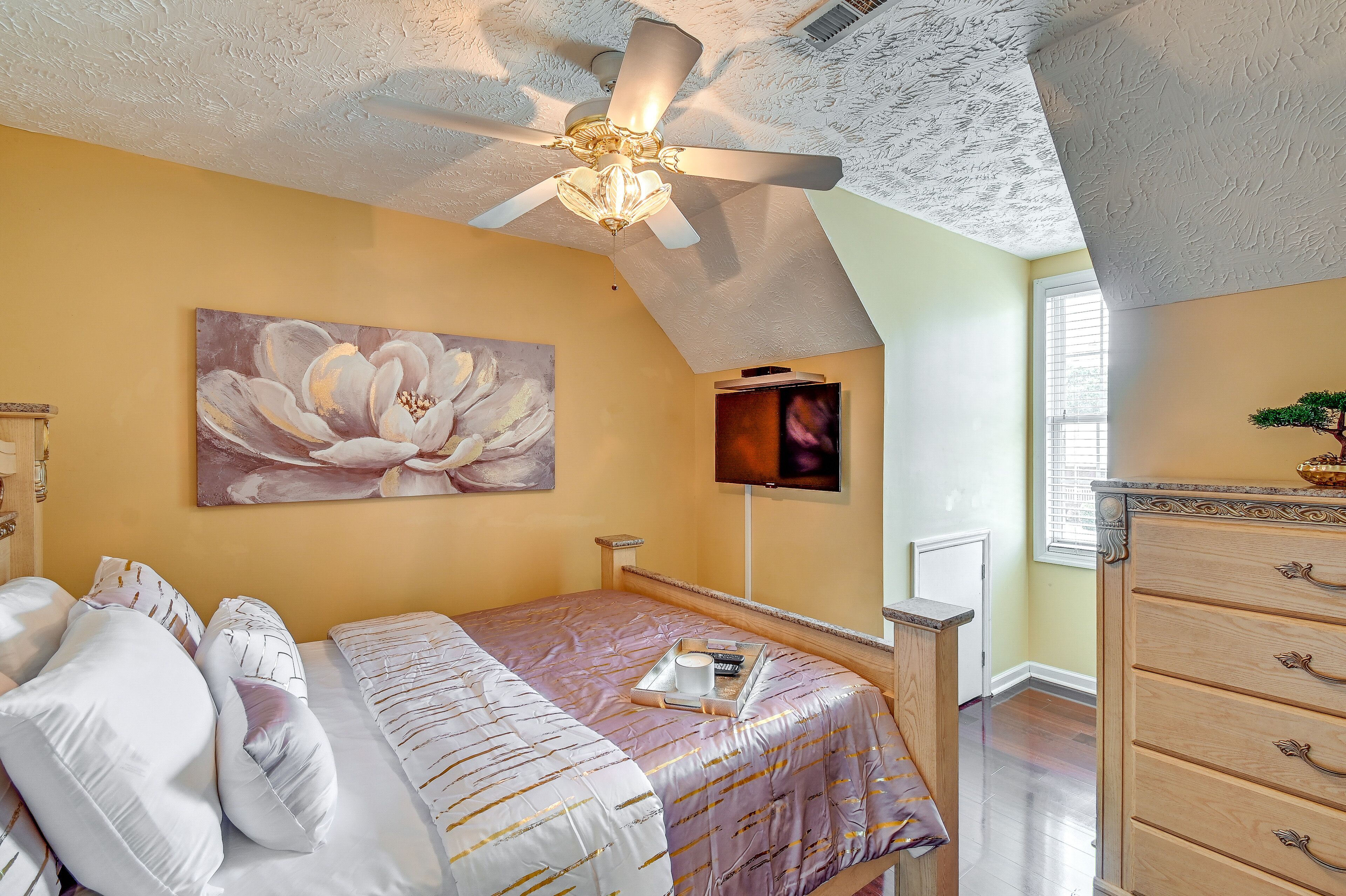 Une chambre à coucher avec un lit, un ventilateur de plafond, un tableau, une commode et une fenêtre.