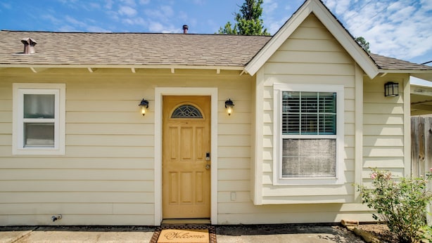 Una casa de un piso con fachada color crema, puerta principal de madera, dos ventanas y un pequeño porche.