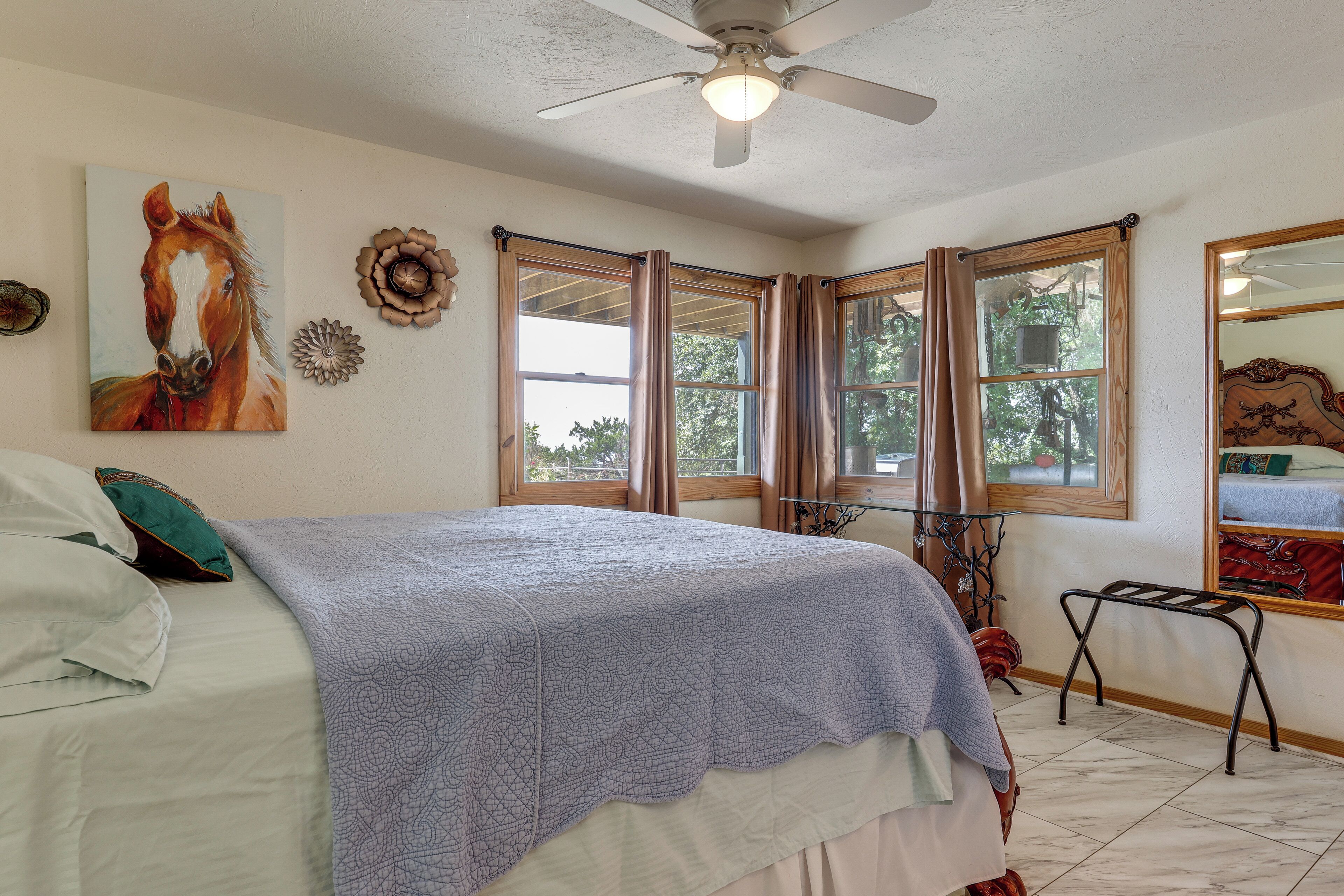 A bedroom with a bed, a ceiling fan, a window with curtains, a painting of a horse, and a mirror.