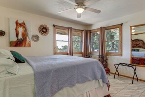 Une chambre à coucher avec un lit, un ventilateur de plafond, une fenêtre avec des rideaux, un tableau représentant un cheval et un miroir.