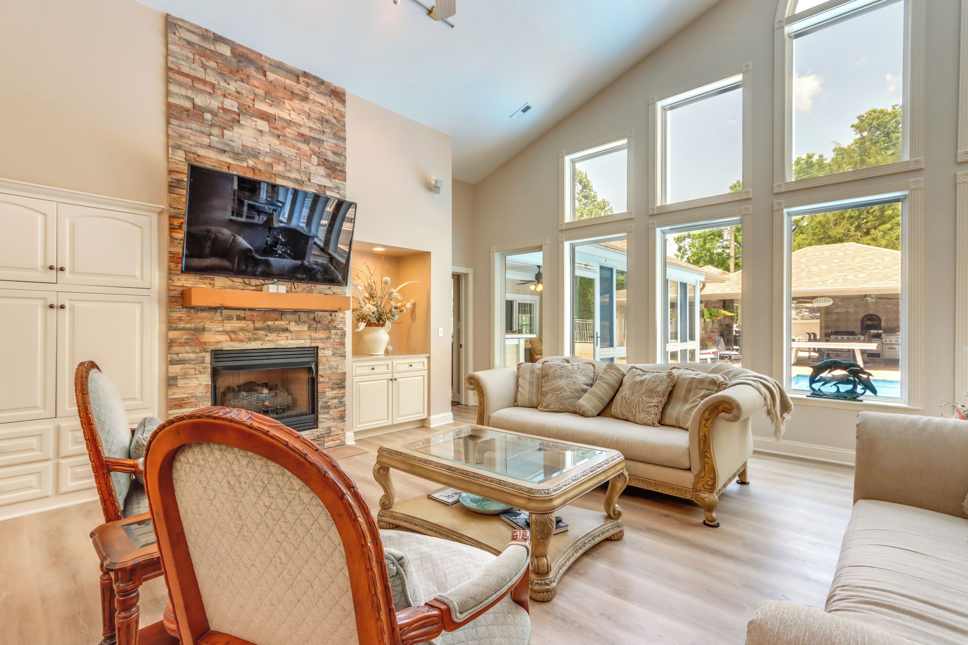 A spacious living room with a fireplace, a large TV mounted on a brick wall, and a view of the outdoors through large windows.