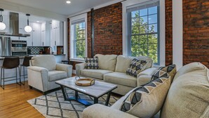 A modern living room with a sofa set, a coffee table, and a kitchen area with white cabinets and a brick wall. - 1 Mi to Falls View Park: Convenient Cohoes Retreat (Cohoes)