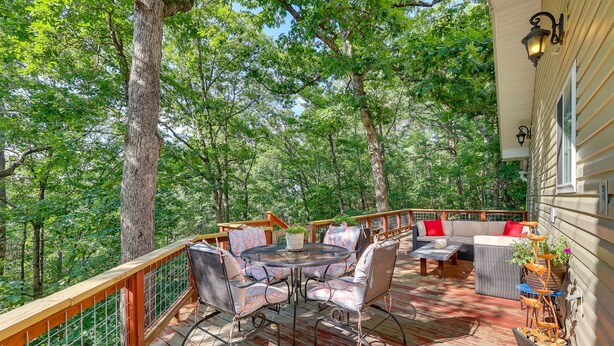 A deck with a round table and chairs, surrounded by a wooden railing and a sofa with red cushions. - Secluded Ozark Mtn Cottage w/ Wraparound Deck (Eureka Springs)