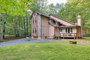 Eine zweigeschossige Holzhütte mit Steinverkleidung, einer Terrasse mit Sitzgelegenheit und einem Wald im Hintergrund.