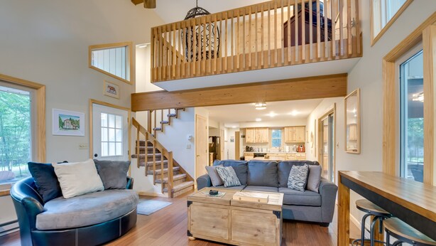 A spacious living area with a curved sofa, a wooden coffee table, and a bar area with stools. A wooden staircase leads to an upper level. - Pocono Lake Retreat w/ Fire Pit: Near Beach! (Pocono Lake)