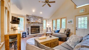 A spacious living room with a stone fireplace, a ceiling fan, and a large window providing natural light. - Pocono Lake Retreat w/ Fire Pit: Near Beach! (Pocono Lake)