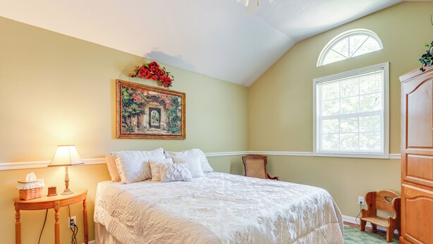 A bedroom with a large bed, a nightstand with a lamp, a chair, a wooden cabinet, and a painting on the wall. - Spacious Family Home Near Lake of the Ozarks! (Camdenton)