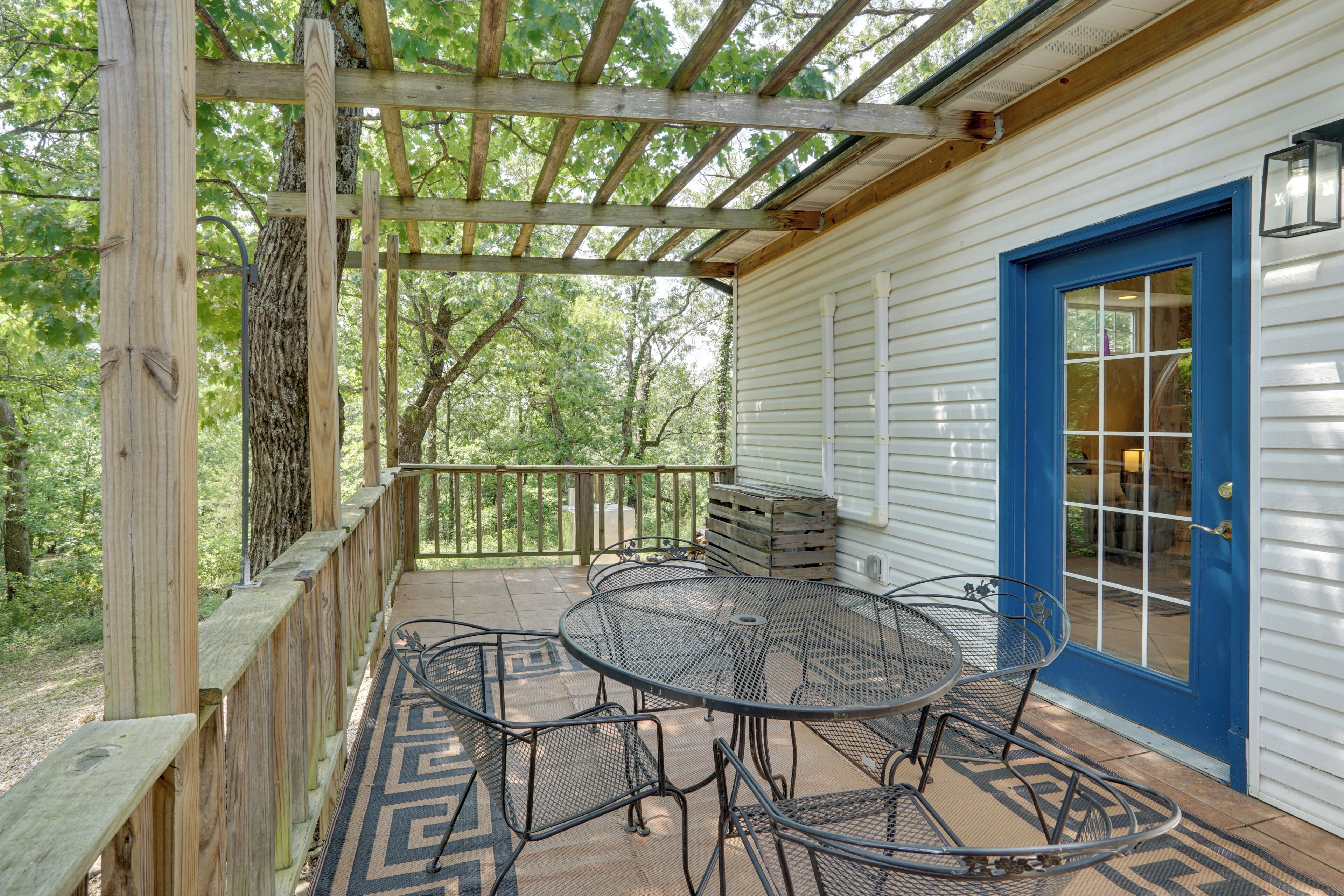 A covered outdoor patio with a table and chairs, a blue door, and a view of the surrounding trees.