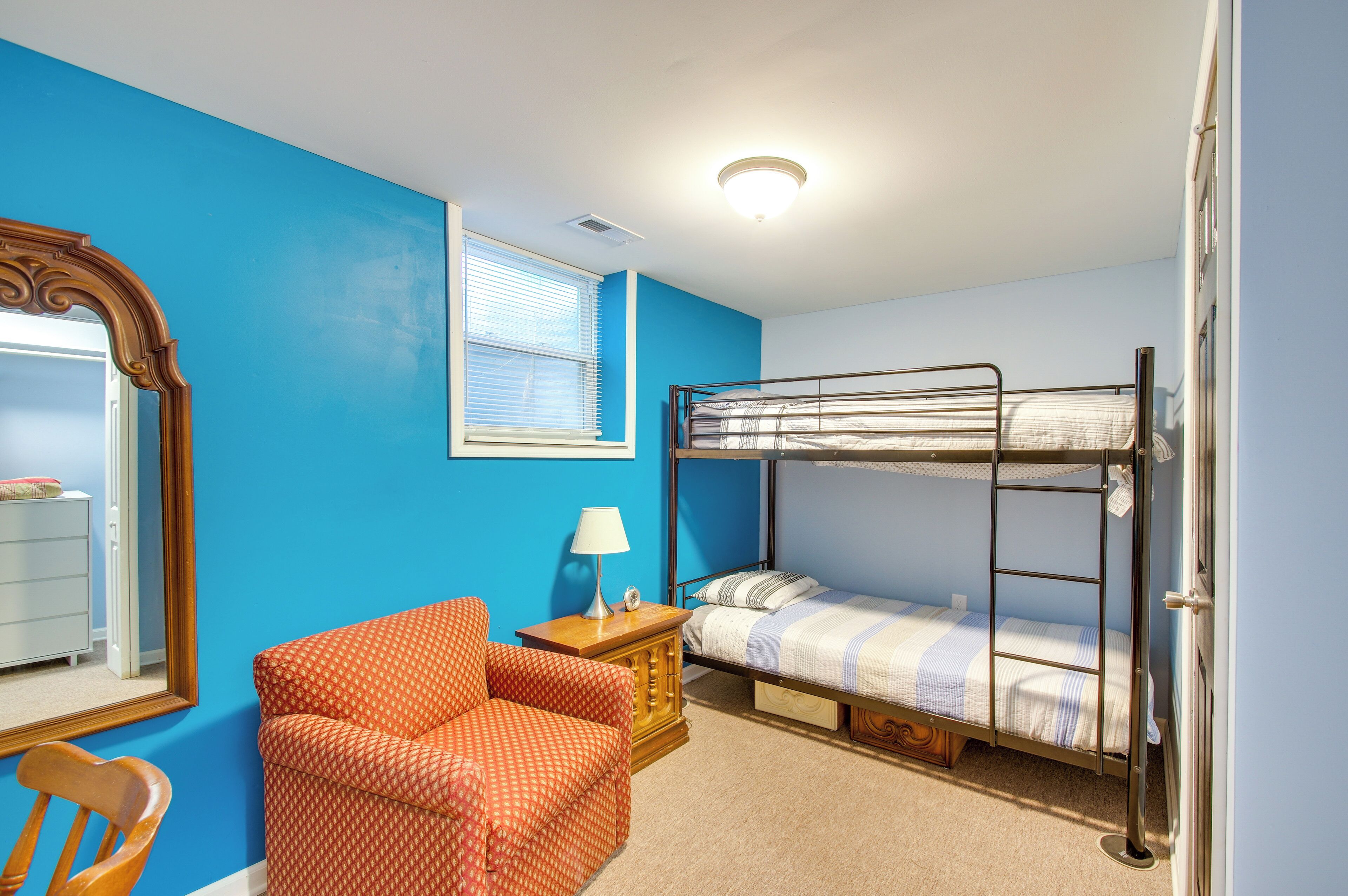 A room with a bunk bed, a chair, a bedside table with a lamp, and a mirror on the wall.