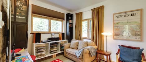 Een gezellige woonkamer met een beige bank, een houten salontafel, een rood-zwart tapijt, een boekenkast, een kaart aan de muur en een raam met gordijnen.