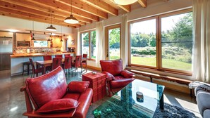 A spacious living area with a wooden ceiling, large windows offering a view of the outdoors, and a modern kitchen in the background. - Acadia Home w/ Rooftop Deck - Close to Trails! (Mount Desert)