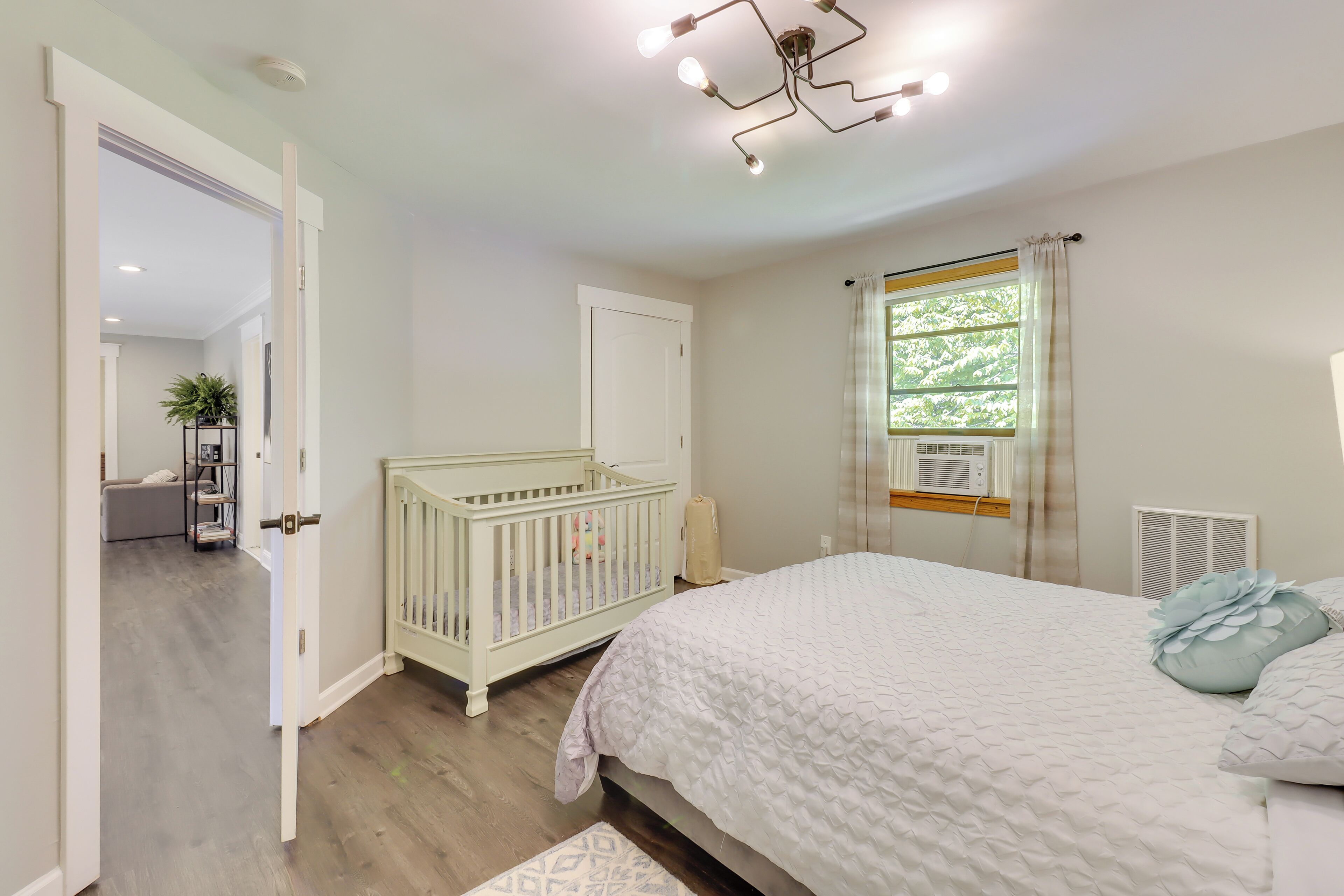 Ein Schlafzimmer mit einem Babybett, einem Bett, einem Fenster mit Vorhängen und einem Flur, der in einen anderen Raum führt.