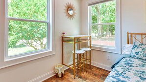 A bedroom with two large windows, a bedside table with a mirror, a bed with floral bedding, and a small stool. - 3 Mi to Dtwn Winston-salem: Studio on Miller Park! (Winston-Salem)