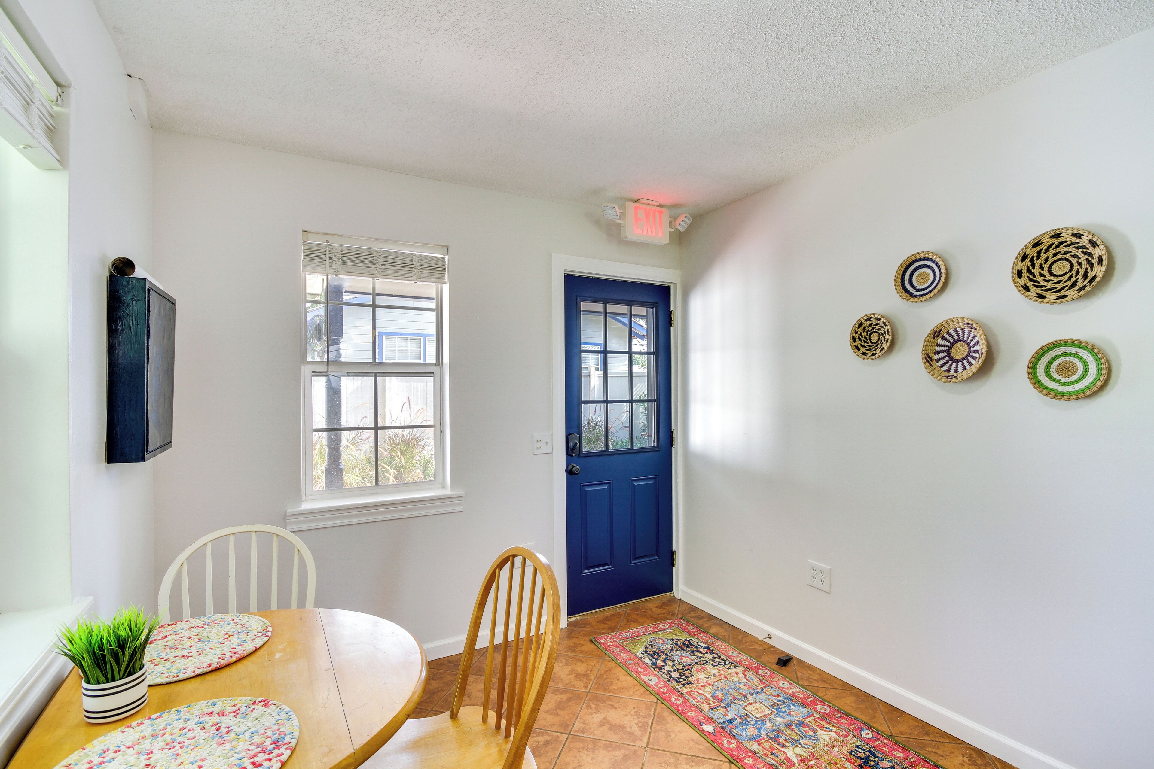 A dining area with a wooden table, chairs, a potted plant, a blue door, and a patterned rug.