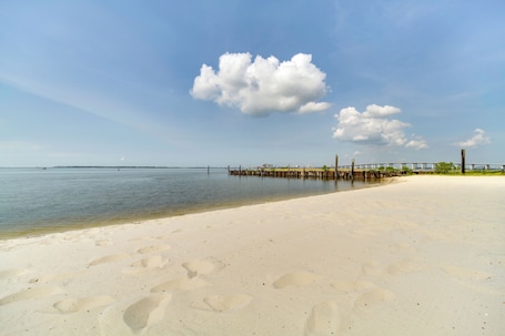 A beach with a wooden pier extending into calm waters, a clear sky, and footprints in the sand.