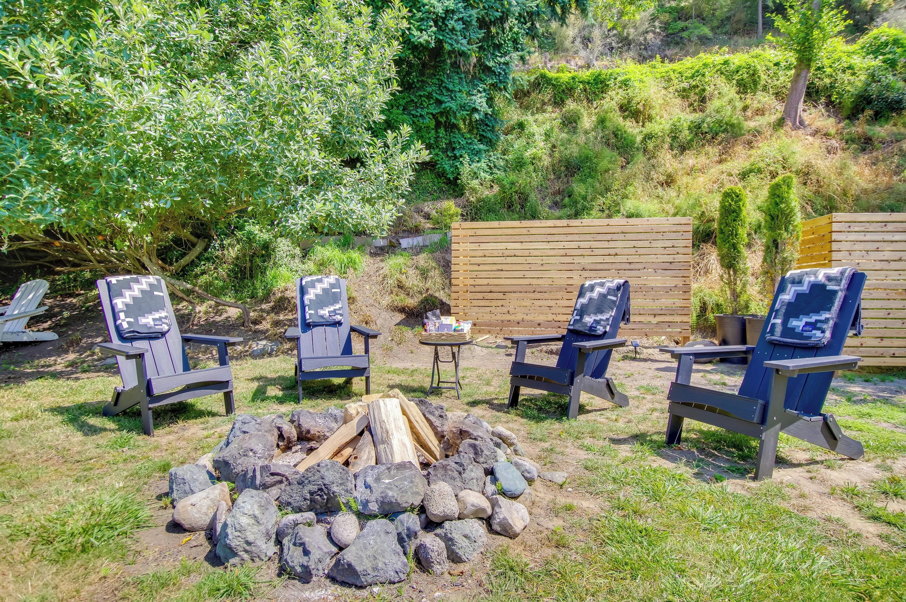 A backyard with a fire pit surrounded by Adirondack chairs, a wooden fence, and lush greenery.