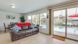 A living room with a brown leather sofa, a red throw blanket, and a view of a swimming pool through large windows. - Riverfront Florida Retreat w/ Pool, Patio & Grill! (Port Charlotte)