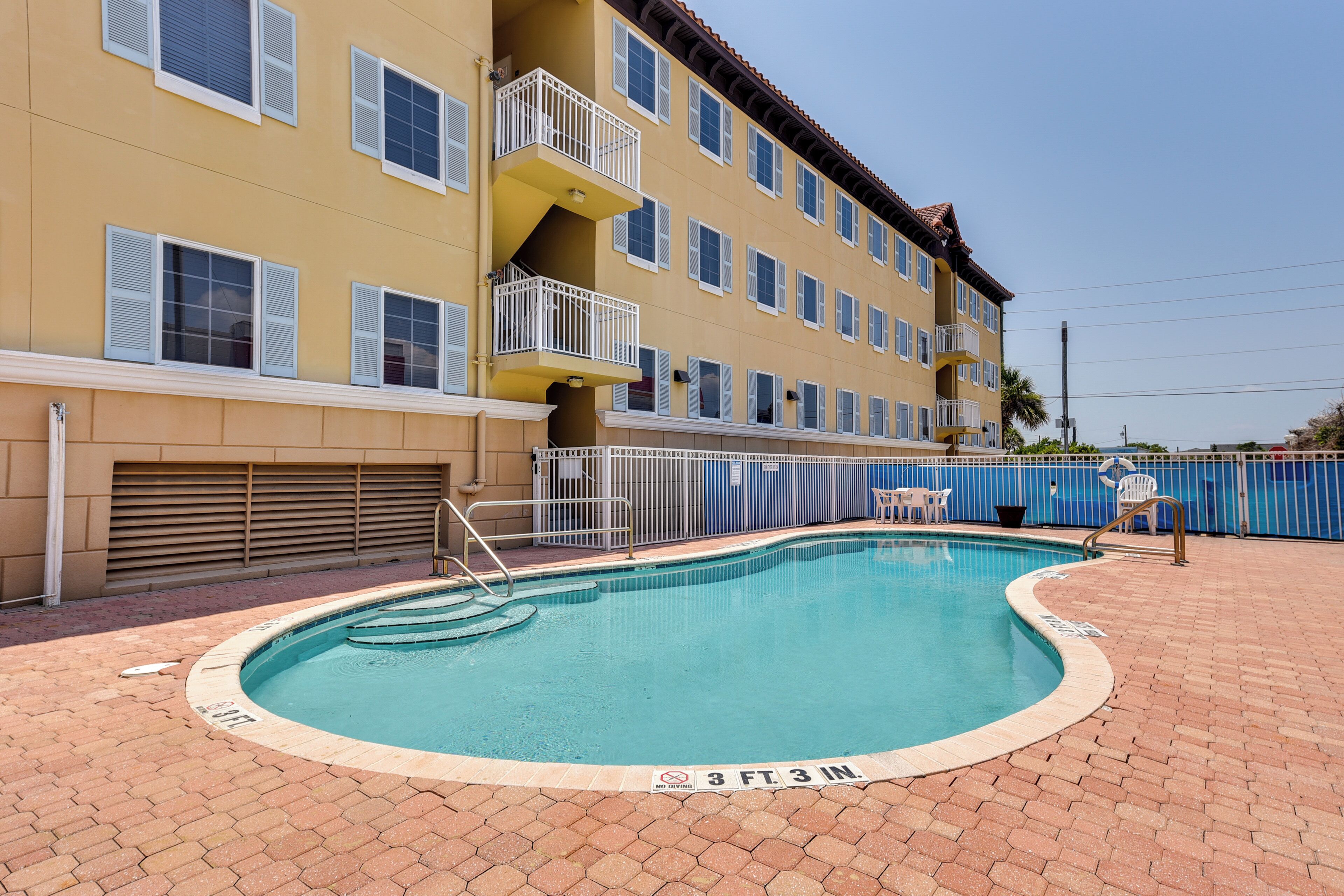 A swimming pool with a 3 ft 3 in. depth marking, surrounded by a brick patio, and adjacent to a multi-story building with balconies and a blue fence.