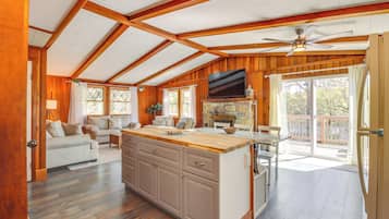 A spacious living area with a wooden ceiling, a fireplace, a TV, and a balcony with a view of the outdoors.