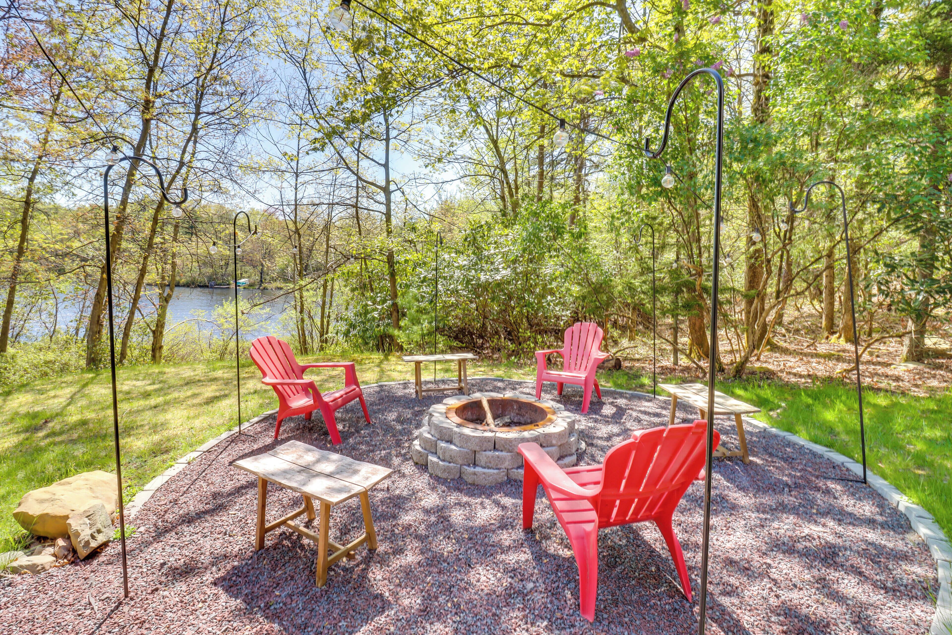 A circular seating area with a fire pit, surrounded by red chairs and wooden tables, set against a backdrop of trees and a lake.