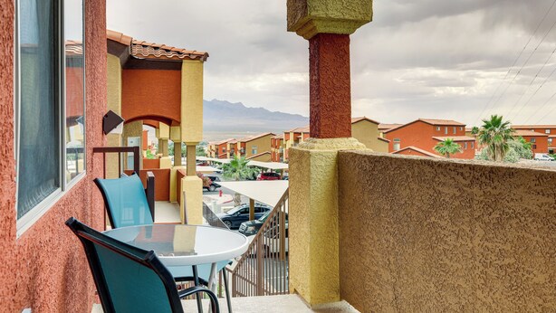 Varanda com mesa e cadeiras, vista para um estacionamento e edifícios com montanhas ao fundo.