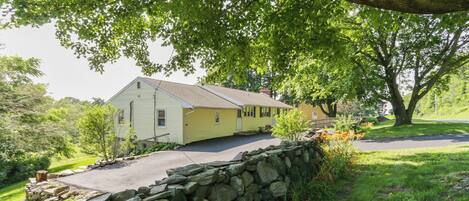 A yellow house with a stone wall in front, surrounded by greenery and trees.