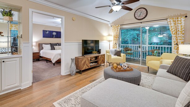 A spacious living room with a sofa, ottoman, and a television. There is a balcony with chairs and a view of trees. - Highend Pawleys Island Condo w/ Porch & Pools! (Pawleys Island)